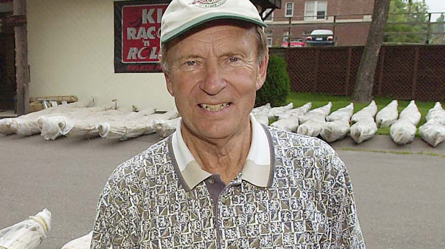 Elderly man in a white cap stands in front of kayaks wrapped in plastic.