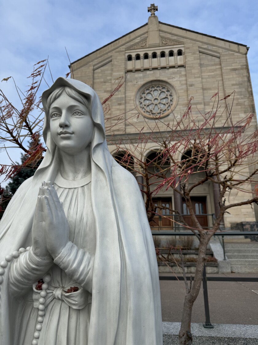 Light stone statue of Mary, mother of Jesus, stands in front of Italianate cathedral. Small trees behind statue indicate fall season.