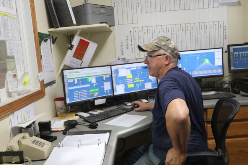 A man in a ball cap sits, watching a processing plant, flanked by large computer screens that monitor feed manufacturing.