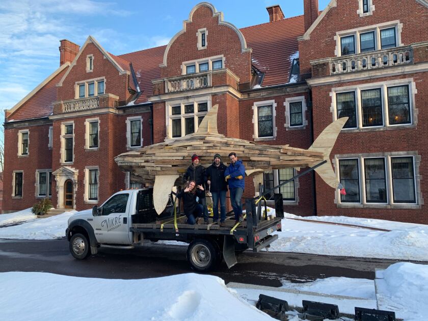 Large wooden shark sculpture is seen atop flatbed truck parked in front of large brick mansion in snowy landscape. Four people pose on truck.