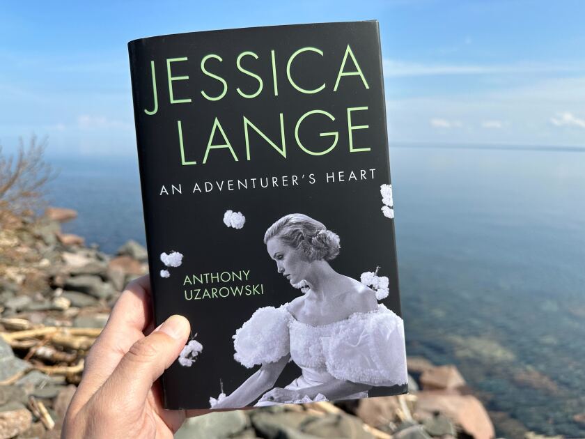White man's hand holds book in front of Lake Superior: "Jessica Lange: An Adventurer's Heart" by Anthony Uzarowski, featuring cover photo of book subject in glamorous white dress.