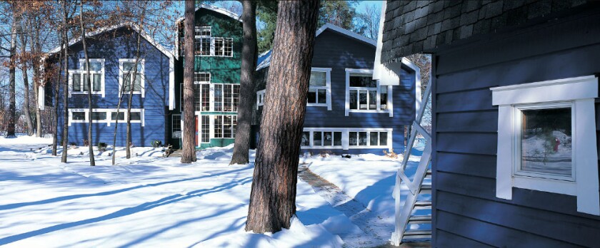 Blue buildings, of modern Scandinavian design and featuring multiple windows, flank a green building in snowy woods with another blue building in foreground right.