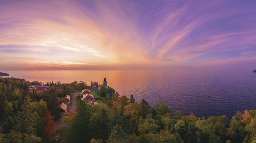 Sun rises over Lake Superior, with color fading from white to a purplish blue, in an aerial photo. Tree-lined shoreline with lighthouse seen in foreground.