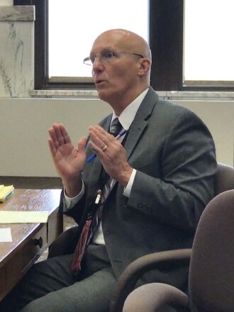 A man seated at a desk gestures while speaking during a meeting
