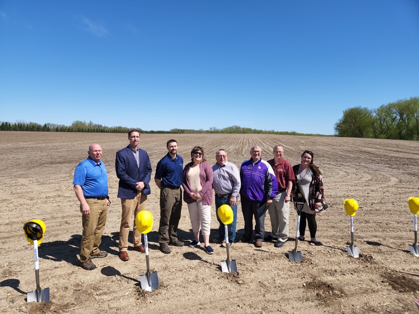 Eight people stand in a dirt covered field behind shovels topped with yellow hard hats.