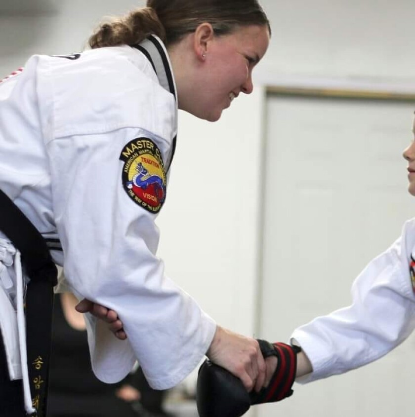 Light-skinned woman, at left, shakes hands with a young person standing largely out of the frame at right. Both wear taekwondo outfits.