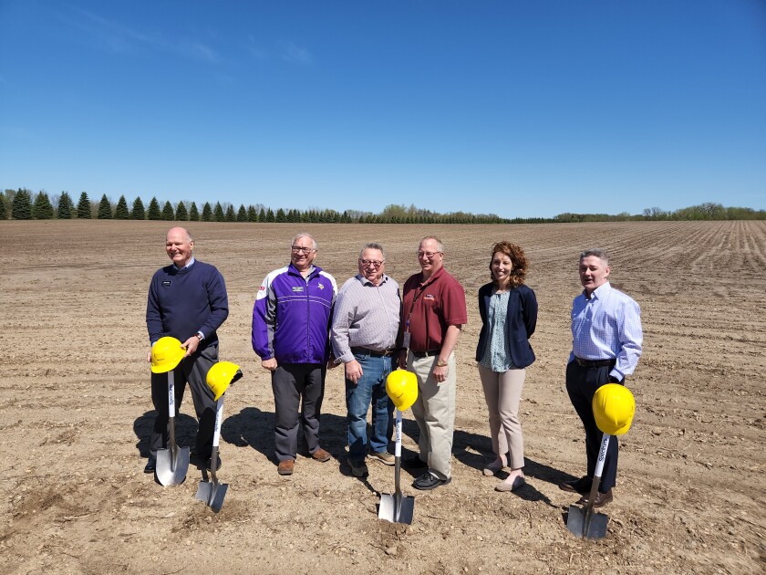 Four Kandiyohi County Commissioners and two representatives from Charter Communications stand in a dirt covered field behind shovels topped with yellow hard hats.