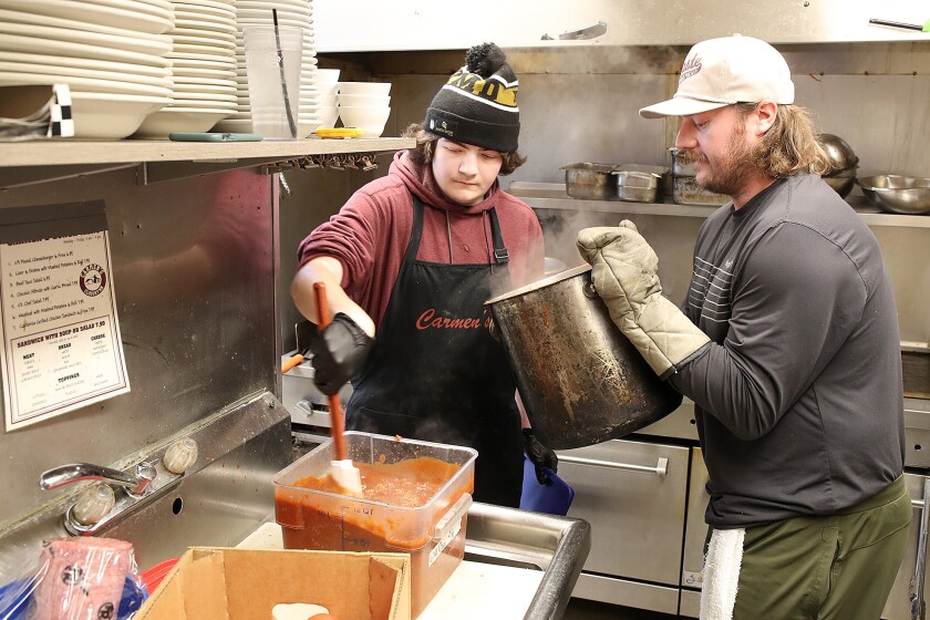 Man on left stirs a container of chili as man on right holds pot of chili in the kitchen of a restaurant.