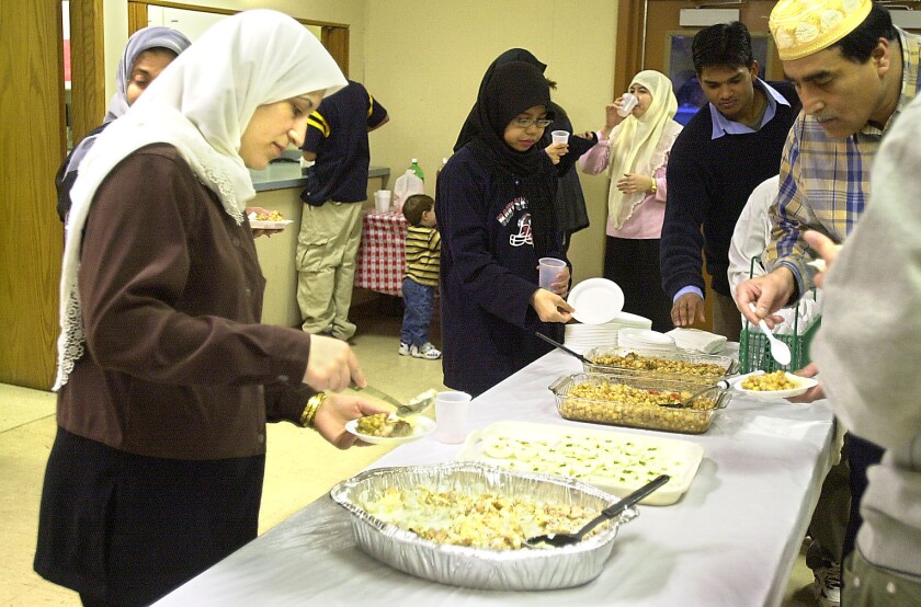 A woman wearing a brown top, black bottoms and a white hijab dishes food onto a small plate, as others hover around a long table with food.