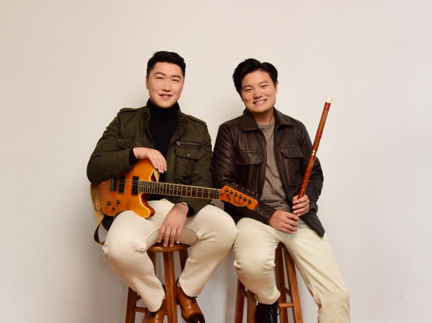 Two men of Asian descent sit smiling on stools in front of white background. Each wears white pants and dark-colored jacket. Man on left holds electric guitar; man on right holds dizi.