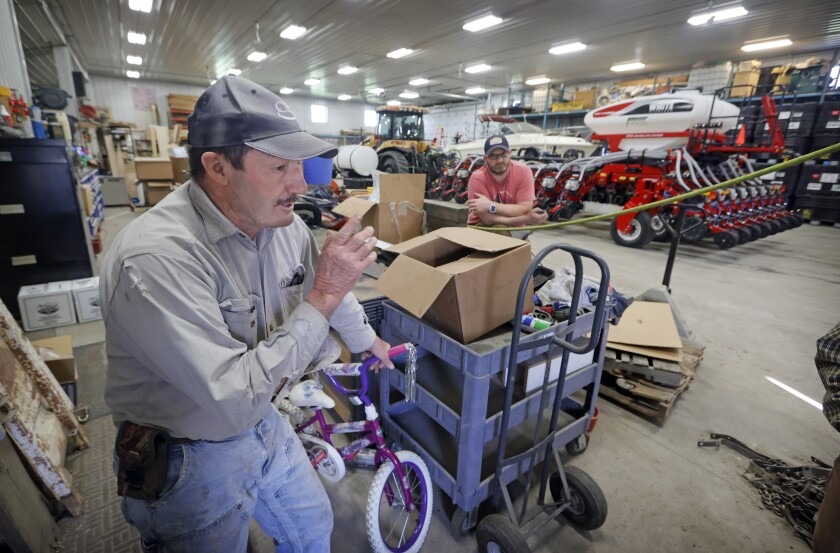 A man in a machinery-filled barn talks while another man leans on a bin in the background.