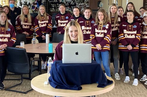 hockey player sits in front of a computer for an interview as teammates gather behind her