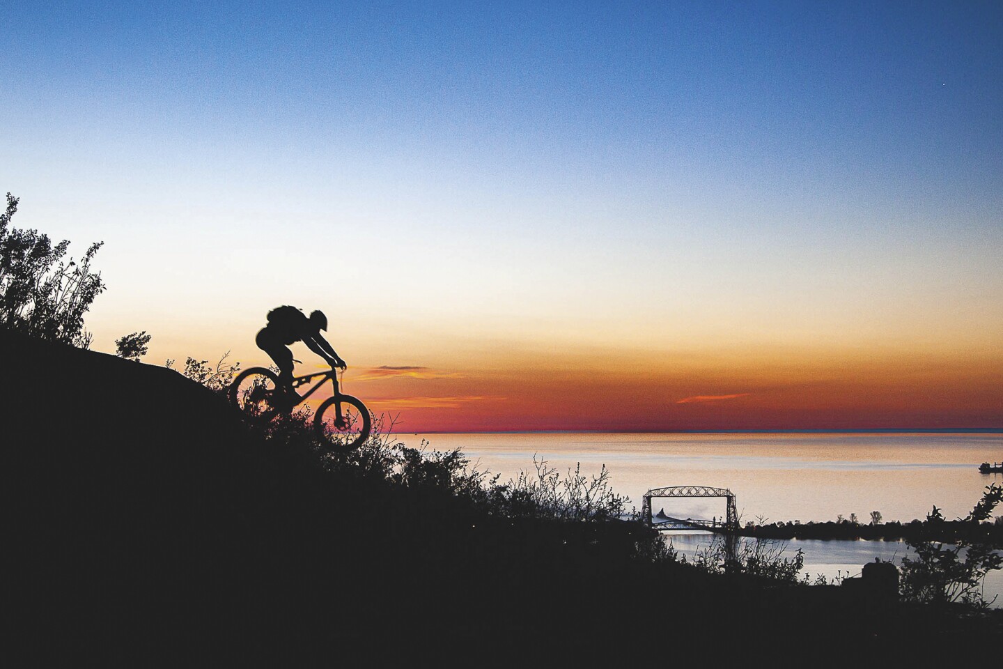 Mountain biker heading downhill is seen in silhouette against a sky darkening over Lake Superior, with Aerial Lift Bridge visible in distance.