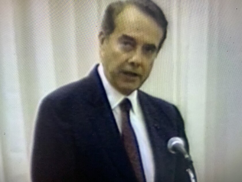 A man in a dark suit speaks in front of a microphone.