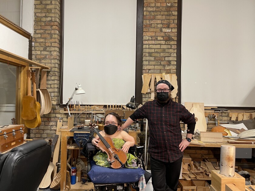 Gaelynn Lea sobre la creación de música para 'Macbeth' de Broadway E! News Reino Unido 2 Una mujer sostiene un violín y se sienta en una silla de ruedas mientras un hombre a su izquierda se apoya en la silla en un estudio lleno de instrumentos musicales.