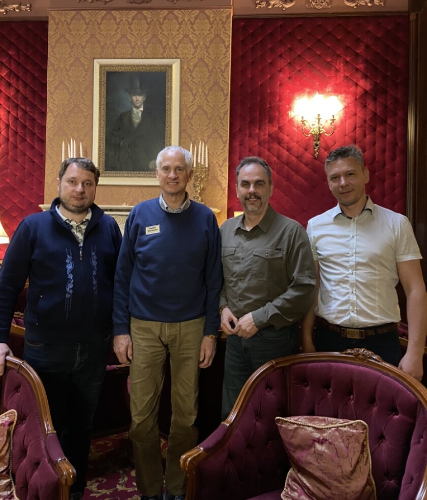 Four men stand in a room with red upholstered chairs and walls.