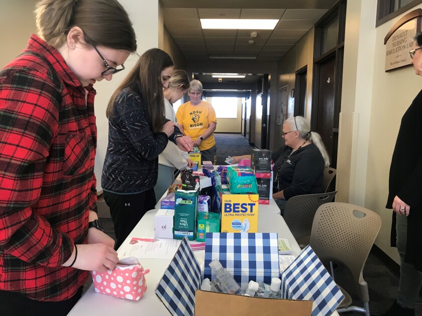 Six people gather around a table and assemble period packs. The table is full of pads, tampons, and hand sanitizer.