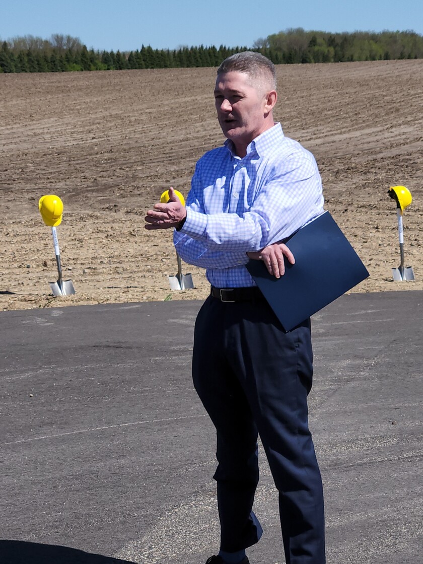 A man stands in front of a dirt covered field addressing attendees of a groundbreaking ceremony.