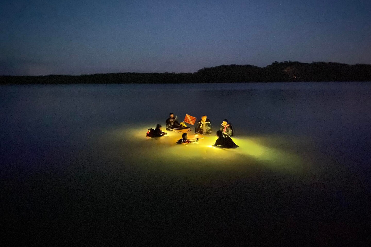 Five divers dressed in scuba gear are lit by an underwater light in the middle of the lake.
