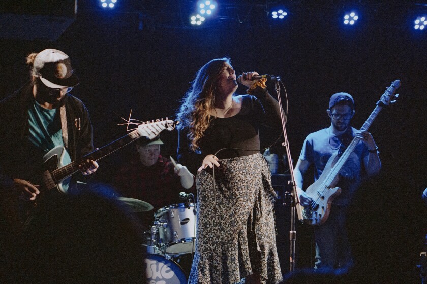 Rock band performing live on a dark stage. A guitarist and bassist flank a singer with long hair wearing a dark blouse and patterned skirt, putting her head back as she sings into a microphone.