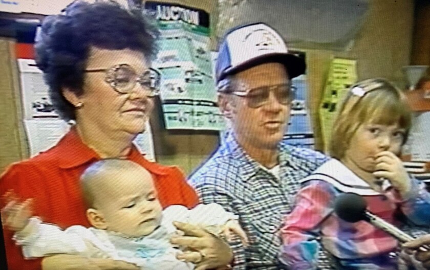 A woman in red holds a baby and a man in flannel holds a small child who sucks her thumb.