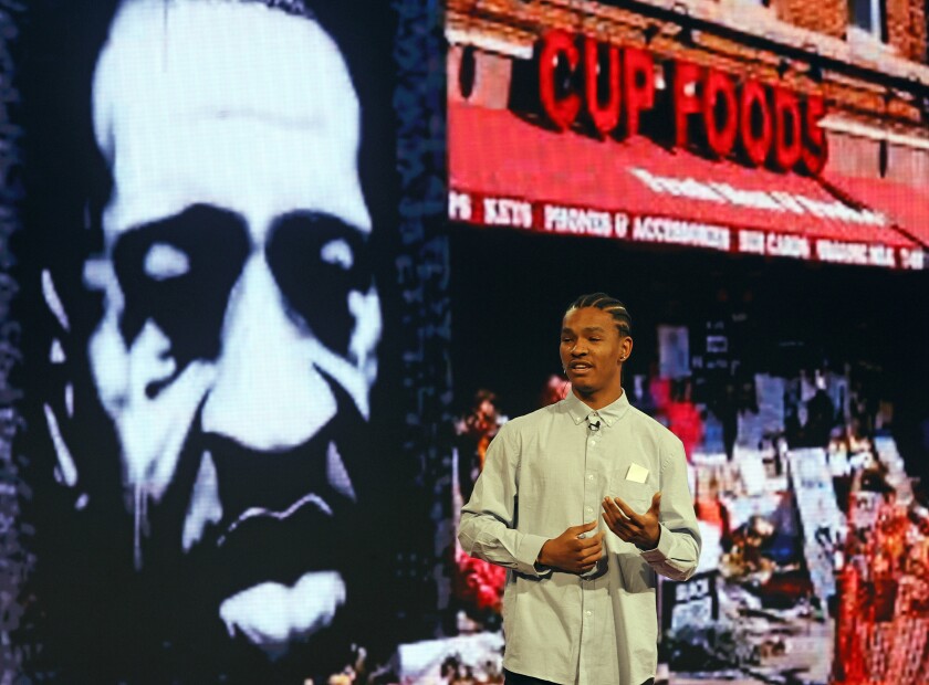 A man in a button-down shirt gestures in front of an image of George Floyd and a photo of the outside of Cup Foods in Minneapolis.