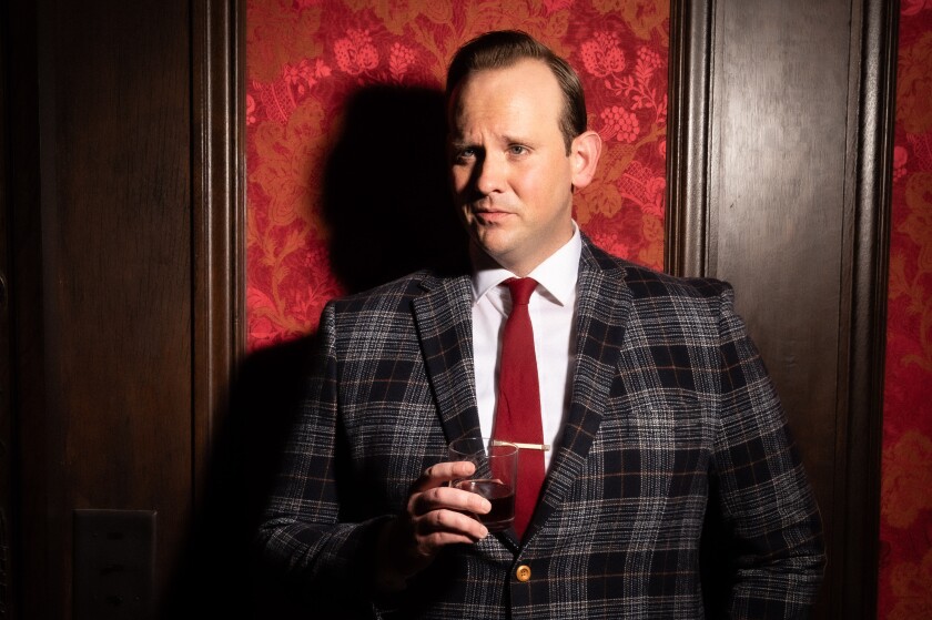Light-skinned man in checkered suit raises eyebrow while holding lowball glass, standing in front of wall finished with polished wood and fine textile.