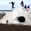 A kid jumping off a snow hill while other kids climb and play on it.