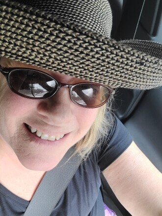 Barbara Sheedy wears sunglasses and a hat as she smiles for a selfie in her car.