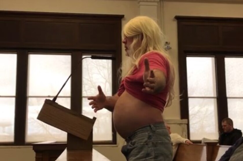 Man wearing long blond wig speaks at a podium