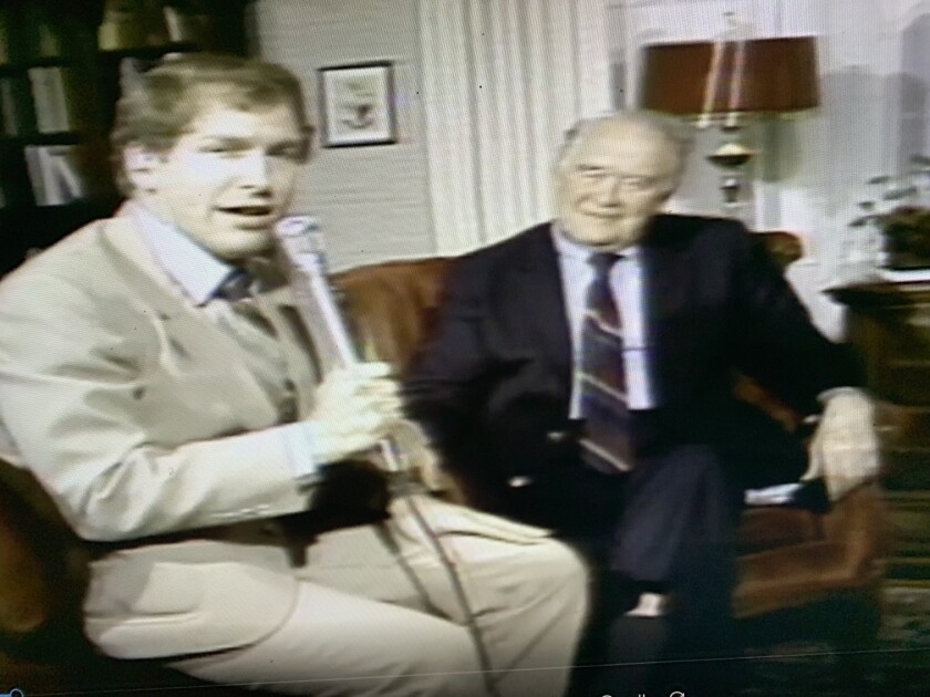 A man in a tan suit talks into a microphone while a man in a dark suit sits next to him in an armchair.