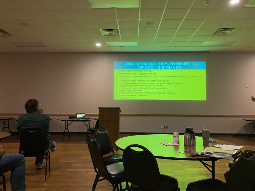 A man wearing a green shirt sits in a chair while he watches a presentation about cover crops on a screen.
