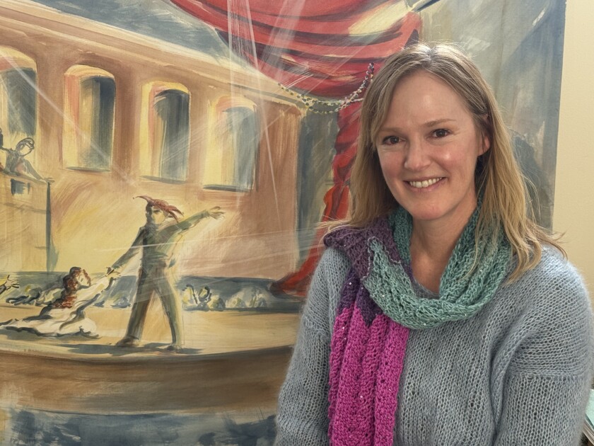 Light-skinned woman with shoulder-length blonde hair smiles while crouching in front of a large painting depicting an Italian figure on stage.