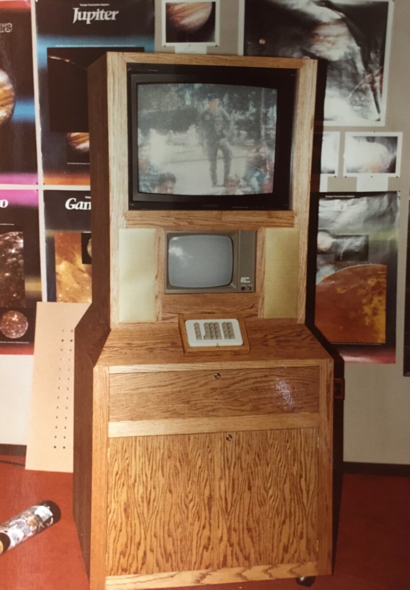 Wood console containing two cathode-ray monitors and a small keypad, displayed in front of posters of planets.