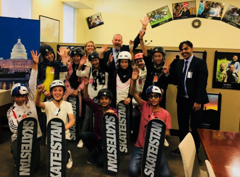 Two adults smile behind two rows of children in helmets holding up skateboards.