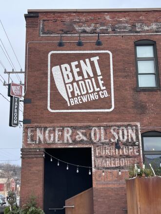 Exterior of Bent Paddle Brewing Co. shows logo painted on the side of a brick building, with faded "Enger & Olson Furniture Warehouse" painted words below