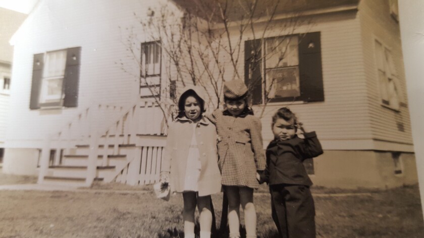 Three young light-skinned children stand holding hands in a black-and-white photo taken circa 1945. Two girls about six years old wear thigh-length coats; a boy about four years old wears a dark suit.
