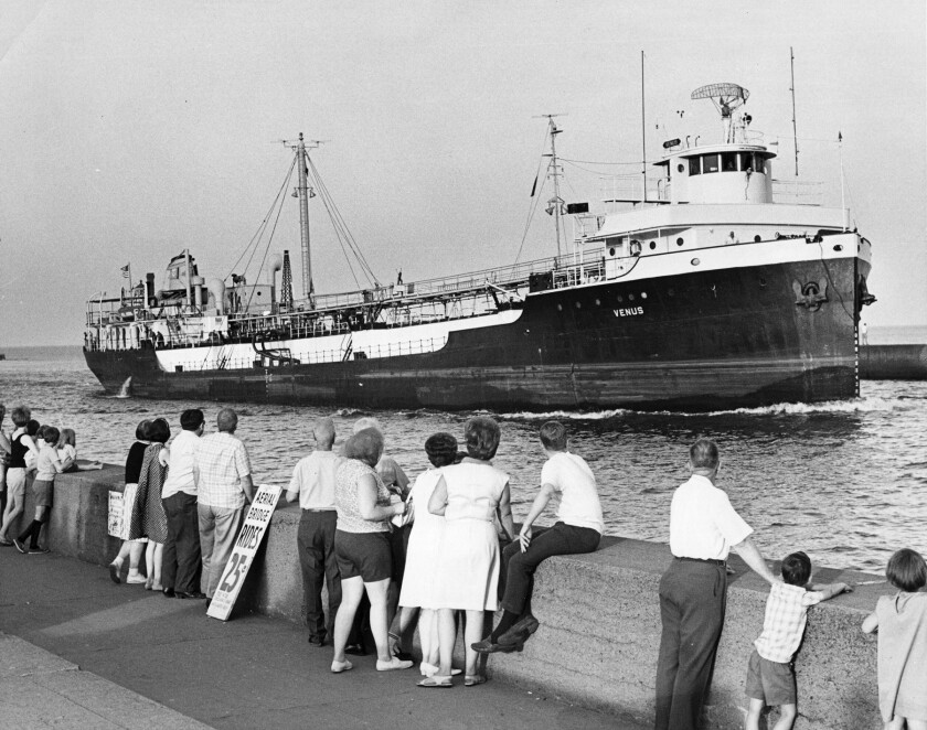 Black-and-white photo of people lining the Duluth Ship Canal's South Pier, with a ship approaching through the canal. A sign leaning against the pier wall reads, "AERIAL BRIDGE RIDES 25 CENTS."
