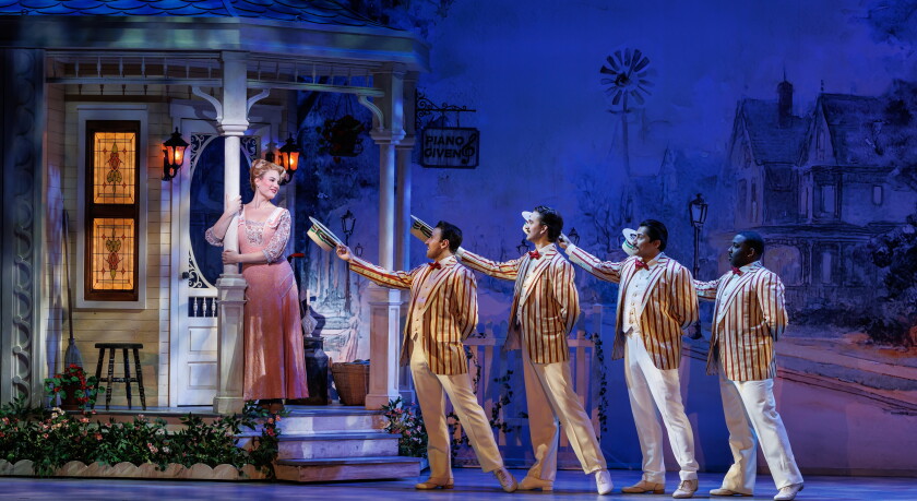Four men wearing pinstripe suit jackets lift boater hats and gesture toward woman wearing pink dress and standing on the porch of Victorian home in a stage set. Other houses, and windmill, are visible on backdrop.