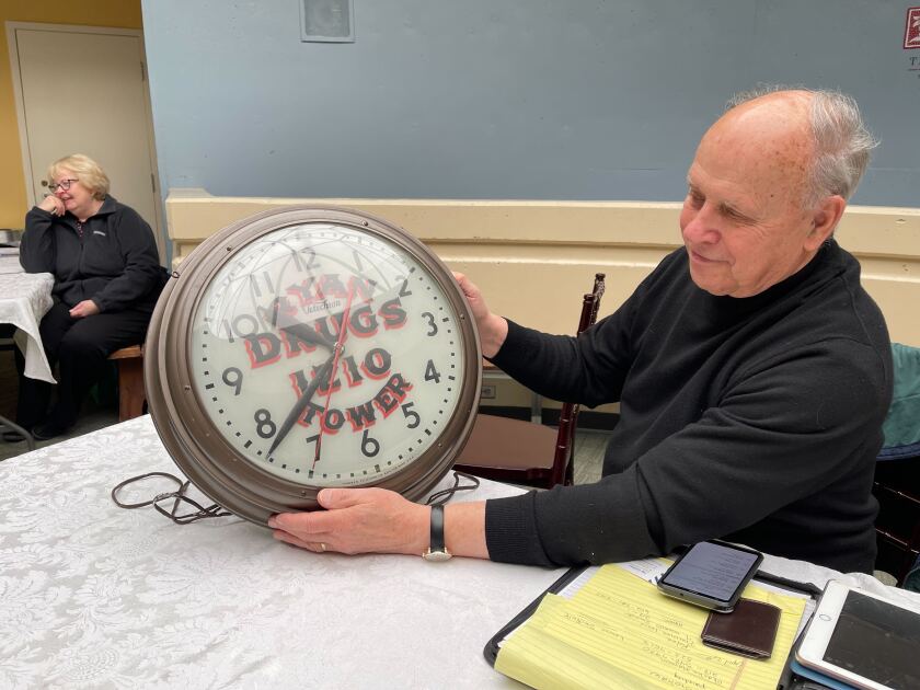 A man holds an antique clock up for display, with the face painted RYAN DRUGS 1210 TOWER. In the background, a woman leans against her elbow and smiles.