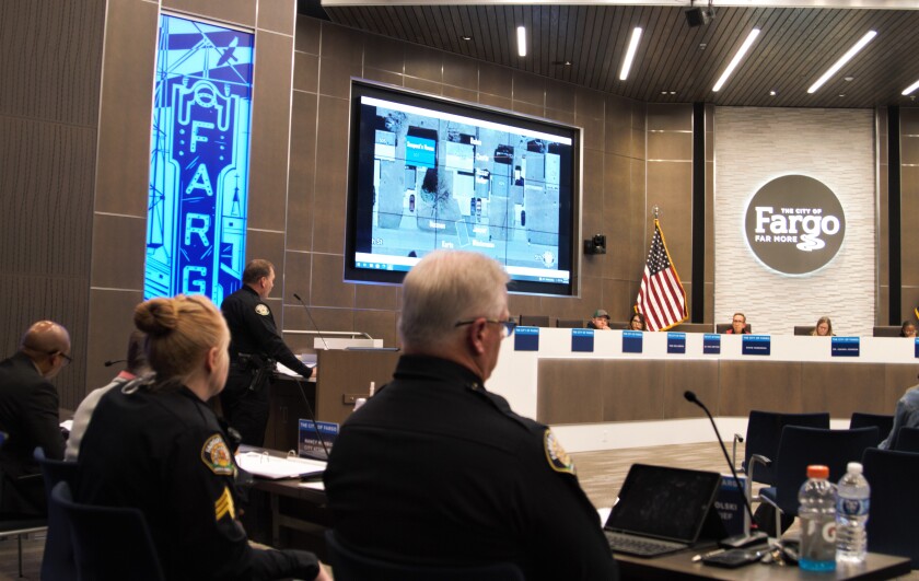(forground) Four people, two of whom are police officers, sit at a table. (midground) Police officer stands at podium. (background) Board members sit at a taller table with a screen on the wall displaying an aerial map.