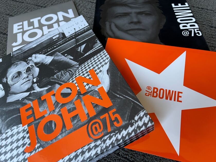 A collection of slipcovers and bold-print envelopes accompanying the books "Elton John @ 75" and "Bowie @ 75"
