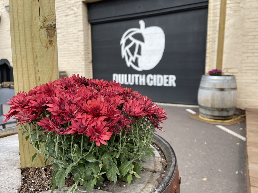 Red flowers are displayed on a barrel in front of garage door bearing Duluth Cider name and logo, slightly out of focus in the background.