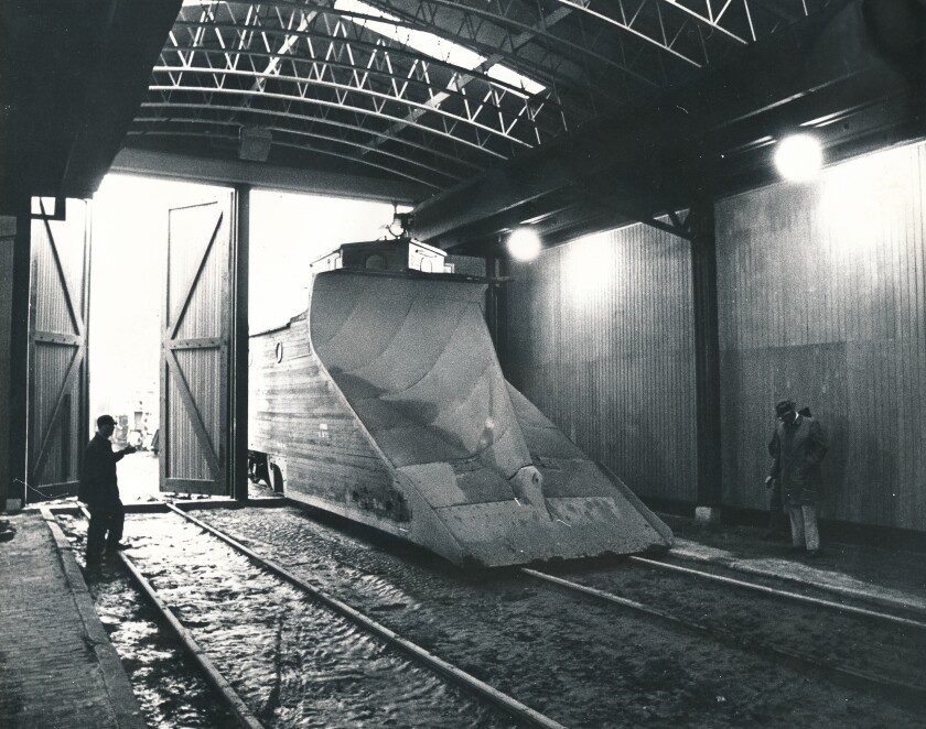 A train snowplow, with large wedge-shaped blade in front, rolls onto an enclosed railroad track in a black-and-white image.