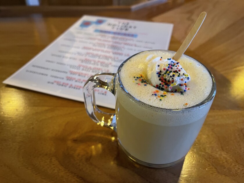 A clear mug is filled with a creamy tan liquid, topped with foam, whipped cream and rainbow sprinkles, sitting on a bar near an out-of-focus menu.