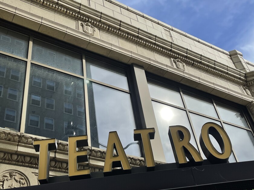 Letters reading TEATRO rise up from a theater marquee, with the reflection of the sun visible in a bank of windows in the background.