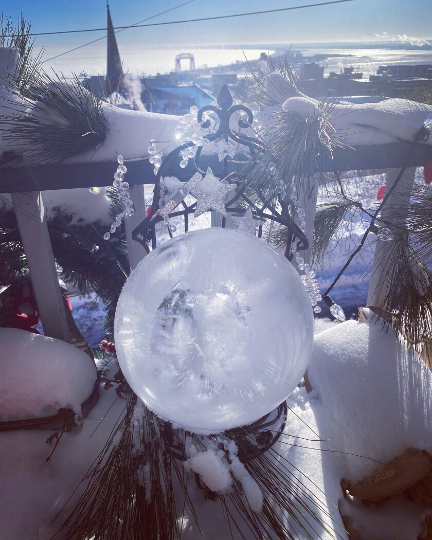 On a snowy deck with a view of Duluth and Lake Superior, a sphere of ice sits in front of pine boughs on a wire-frame chair.