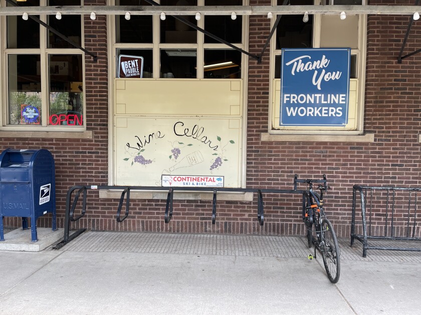 A wide black bike rack, with a single bicycle locked to it, stands against a brick wall with a mailbox to the left and another rack to the right. A sign on a window reads, "Thank you frontline workers."