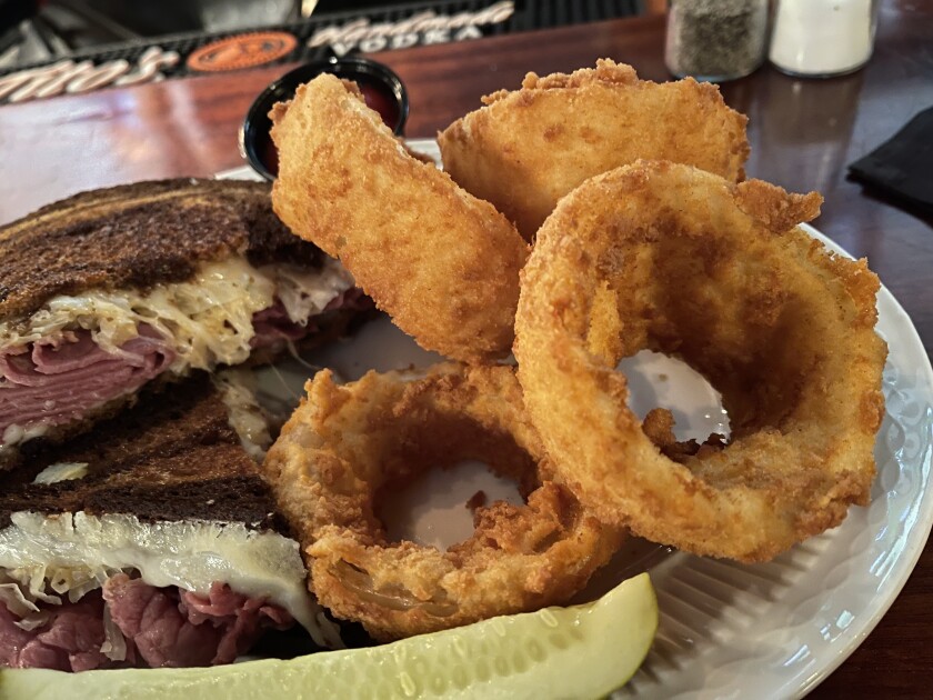 Four thickly breaded onion rings sit on a plate shared with a slice of pickle and a Reuben sandwich