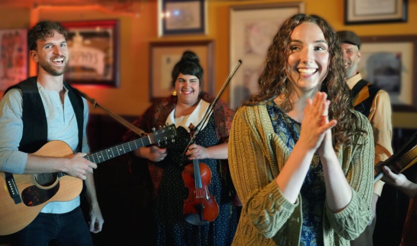 Four people dressed in cozy multilayered garments smile and hold wooden string instruments in a pub-like setting.